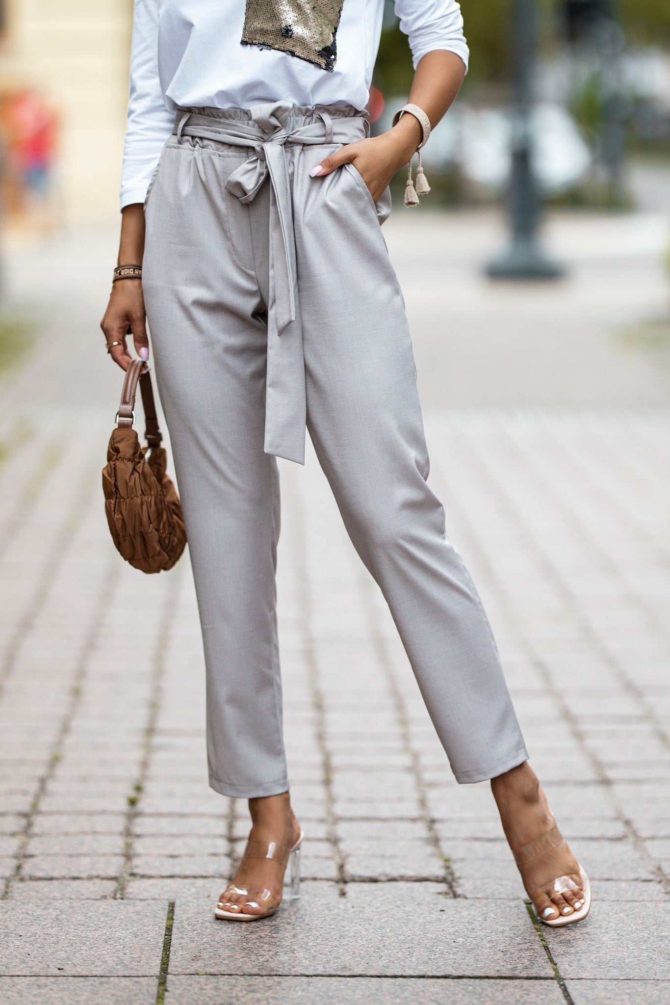 Person wearing light gray pants with a belt, white shirt, and clear heels on a blurred street background.