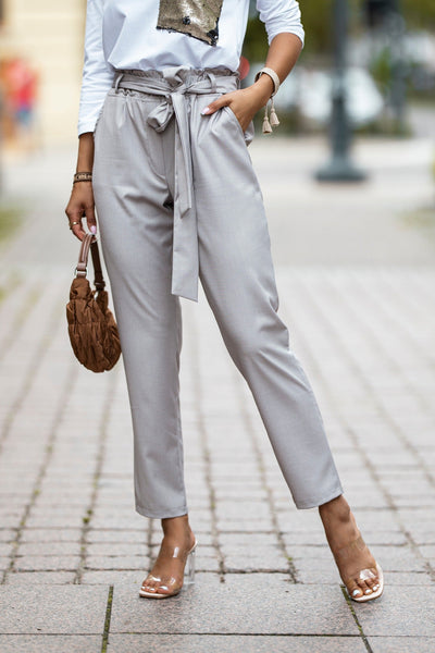Person wearing light gray pants with a belt, white shirt, and clear heels on a blurred street background.