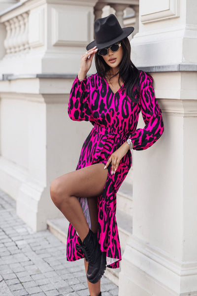 Woman in a pink leopard print dress and black hat leaning against a white wall.