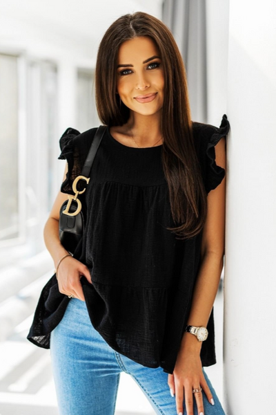 Woman wearing a black top with ruffled sleeves and light blue jeans, standing against a white wall.