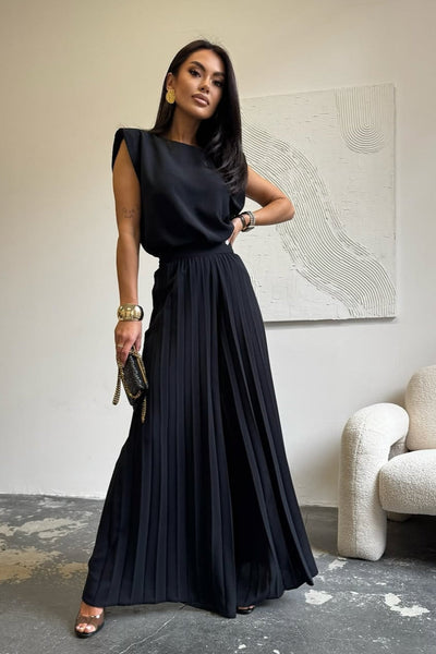 Woman in a black dress standing in a room with a white wall and a chair.